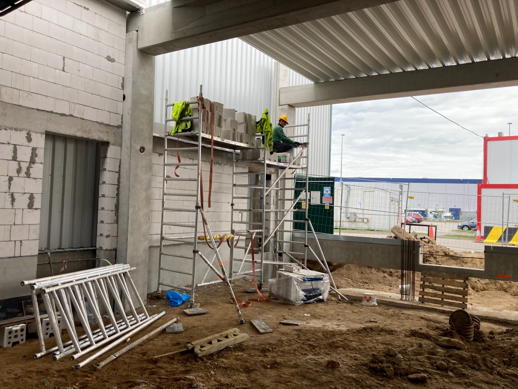 Pracownik w żółtym kasku siedzi na rusztowaniu, układając białe bloczki na budowie hali z widocznymi elementami żelbetowymi i murowanymi ścianami, przy ziemi leżą złożone rusztowania.