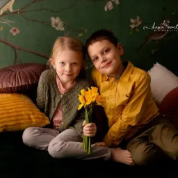 Portret rodzeństwa: uśmiechnięta dziewczynka w zielonym swetrze i chłopiec w żółtej koszuli siedzą na kanapie z poduszkami, trzymając bukiet żółtych narcyzów na tle ciemnozielonej tapety w kwiaty...