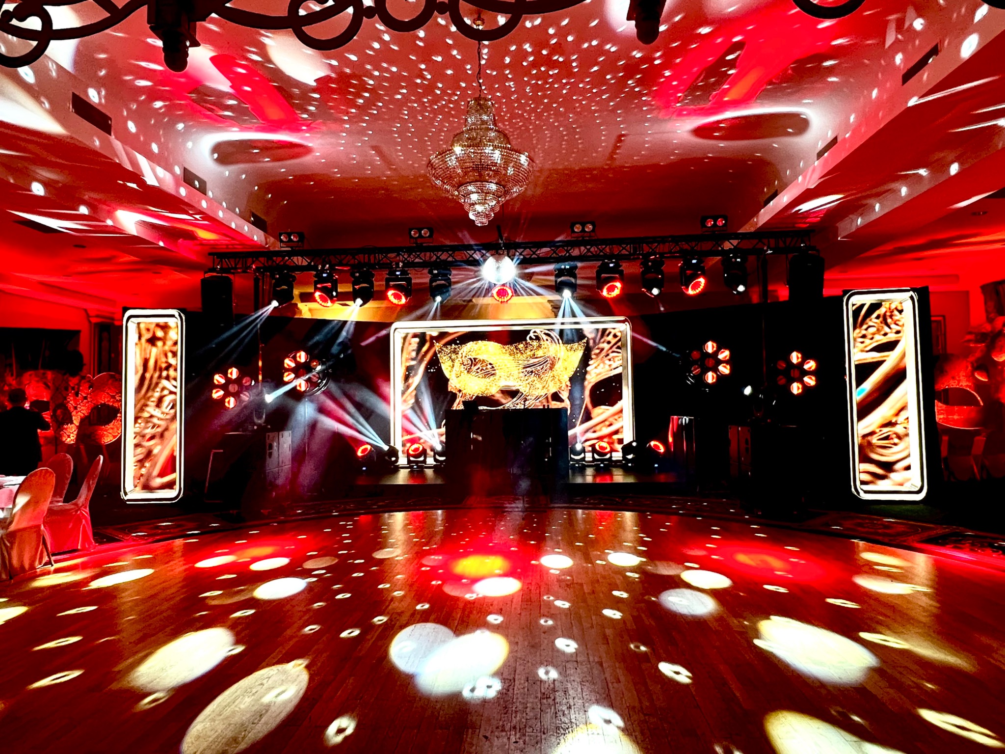 Sala balowa z parkietem oświetlonym reflektorami, scena z ekranem i dekoracjami świetlnymi, kryształowy żyrandol, czerwona kolorystyka.