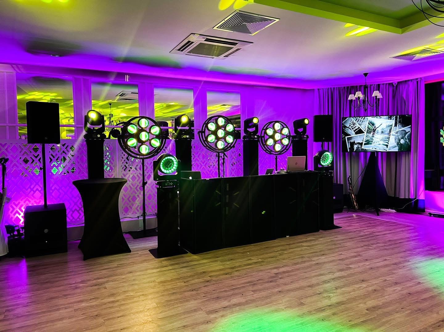 Sala eventowa z oświetleniem scenicznym, konsoletą DJ-ską i ekranem wyświetlającym czarno-białe zdjęcia, oświetlona fioletowym i zielonym światłem.