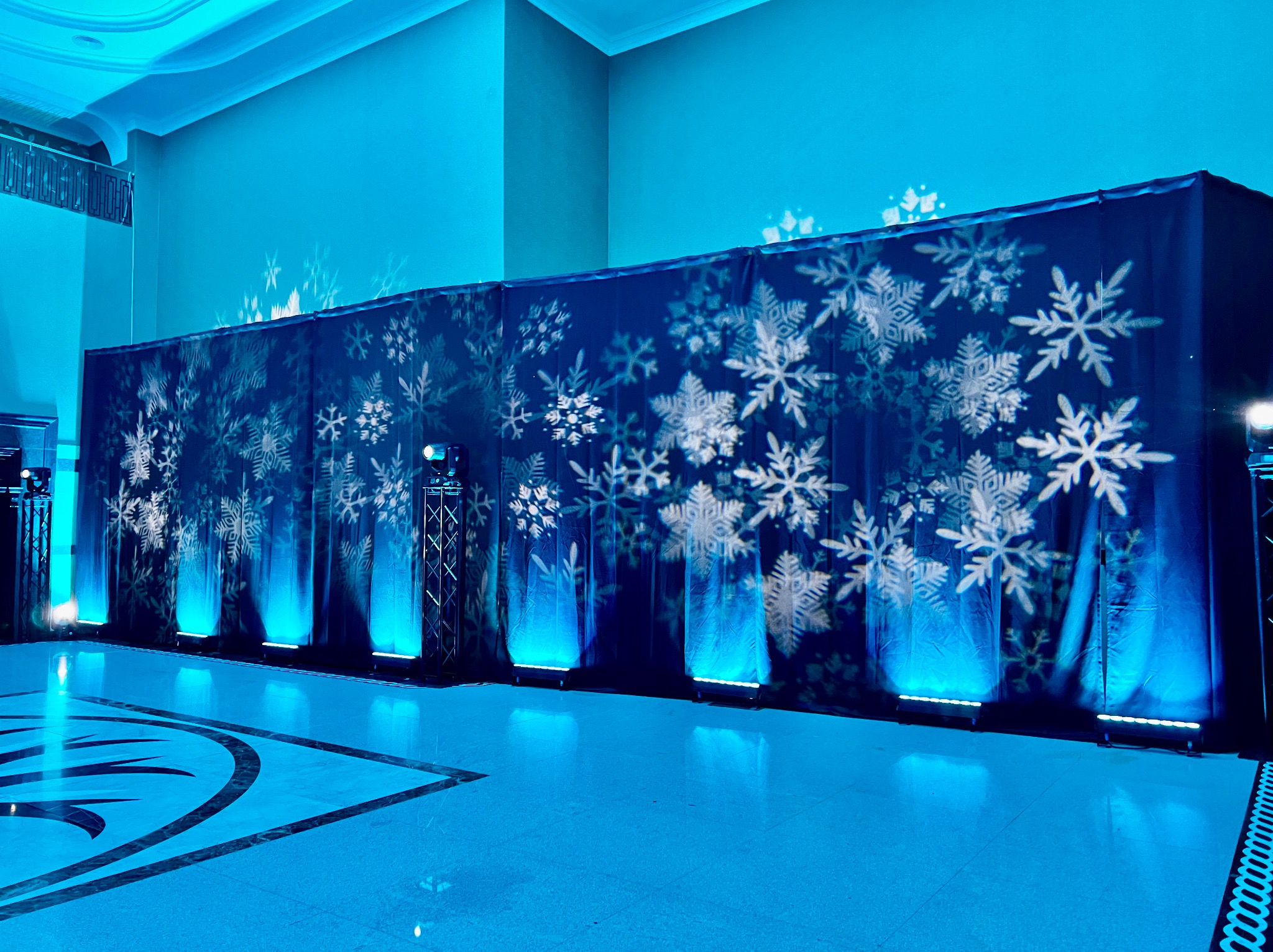 Dekoracja sali balowej w zimowej stylistyce: ciemnoniebieskie zasłony z białymi płatkami śniegu, oświetlone niebieskimi reflektorami podłogowymi, tworzące chłodny, uroczysty nastrój.
