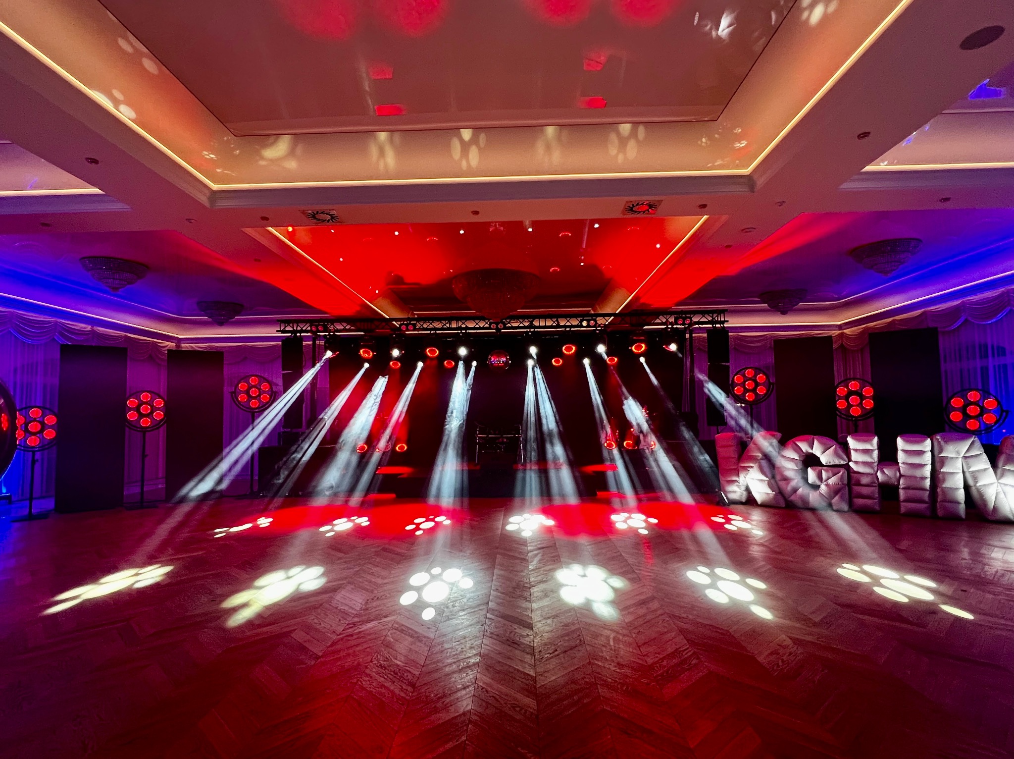 Scena oświetlona czerwonym i białym światłem, z dmuchanymi białymi literami z przodu, parkiet w jodełkę odbija światła, w tle widoczne oświetlenie sceniczne i kurtyny.