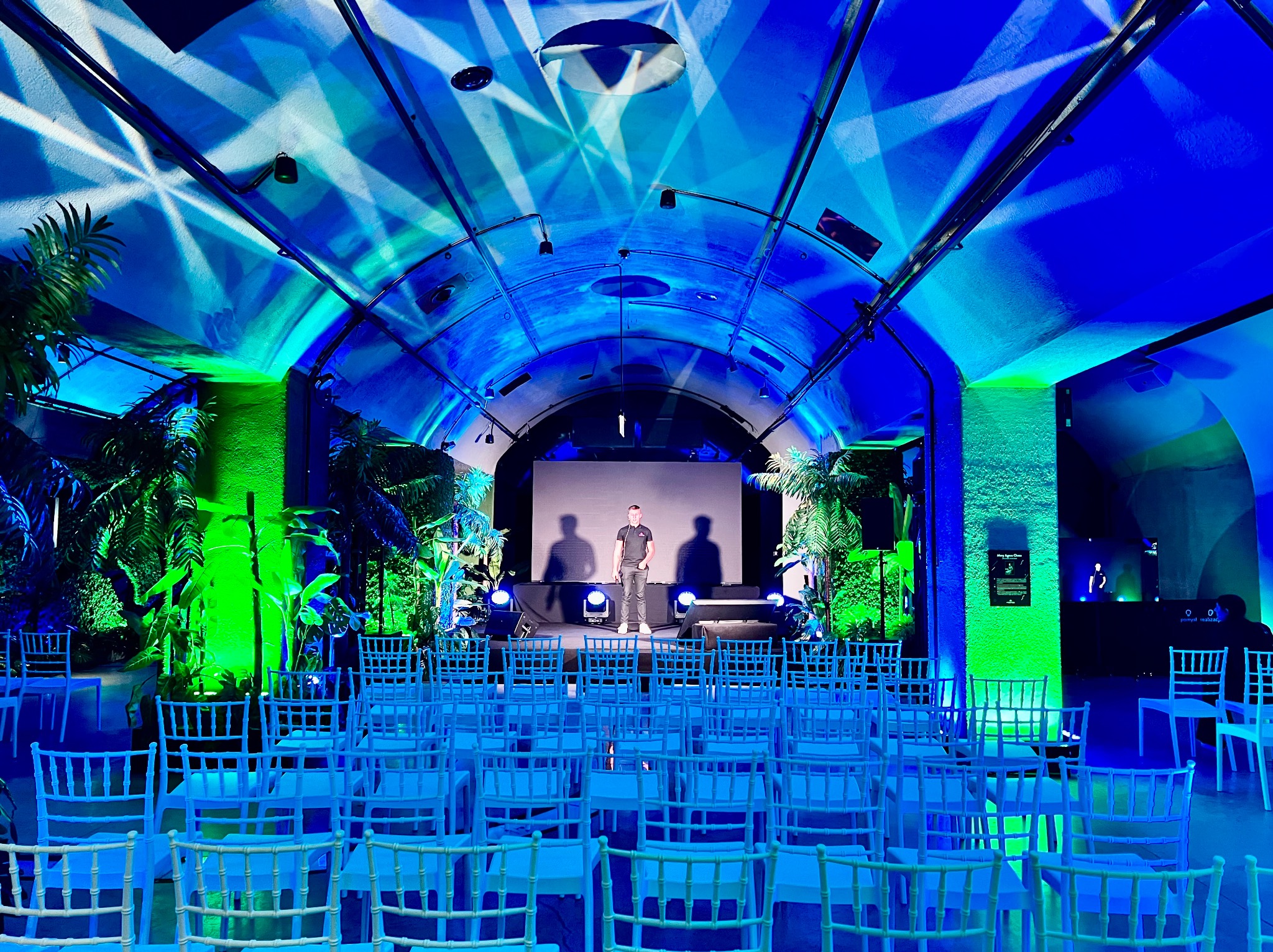 Sala konferencyjna z rzędami białych krzeseł, oświetlona niebieskim i zielonym światłem, z osobą stojącą na scenie przed dużym ekranem.