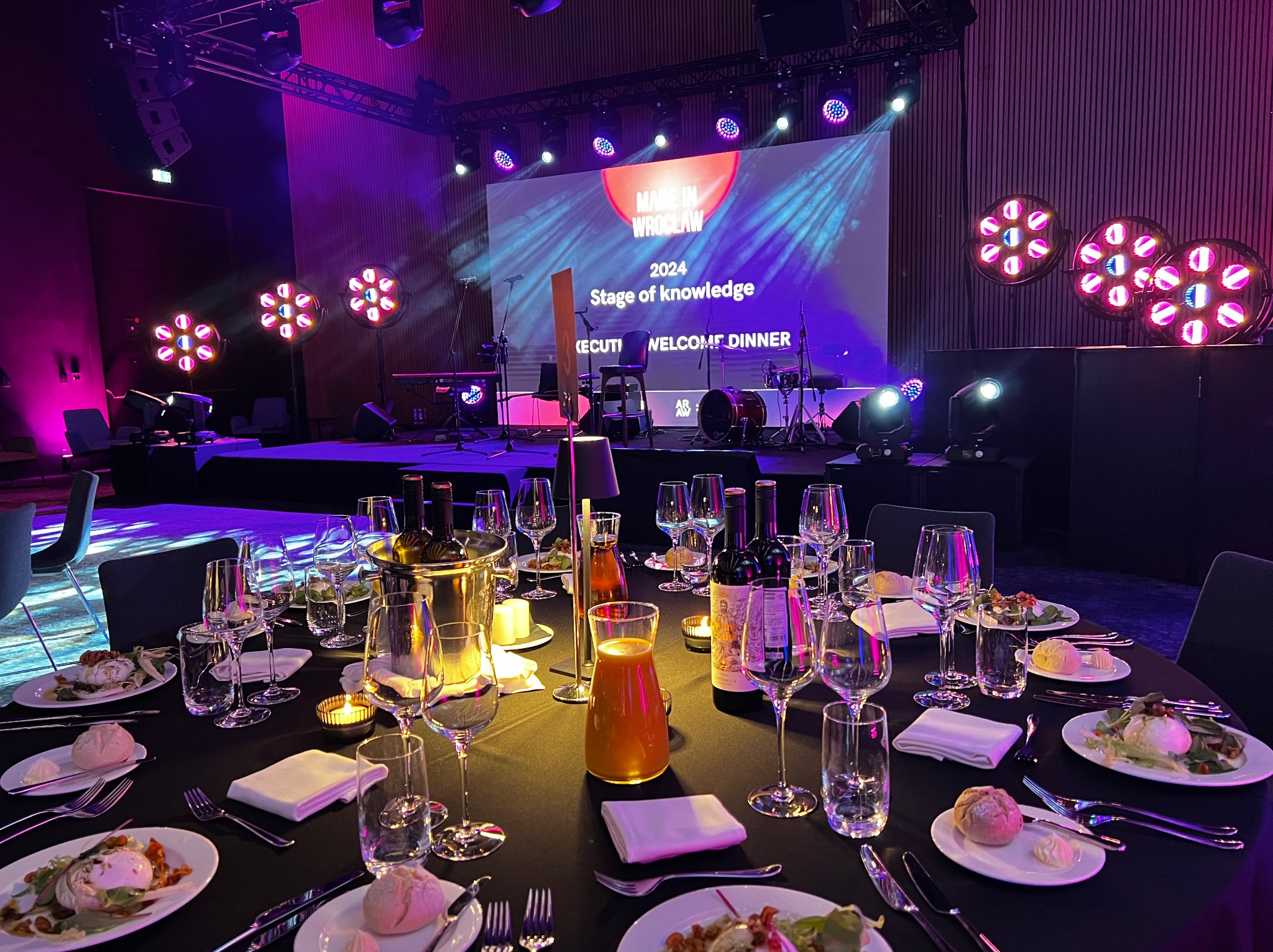 Uroczysta kolacja powitalna z elegancko nakrytymi stołami, oświetlona sceną i ekranem z napisem 'Made in Wrocław 2024 Stage of Knowledge Welcome Dinner'.