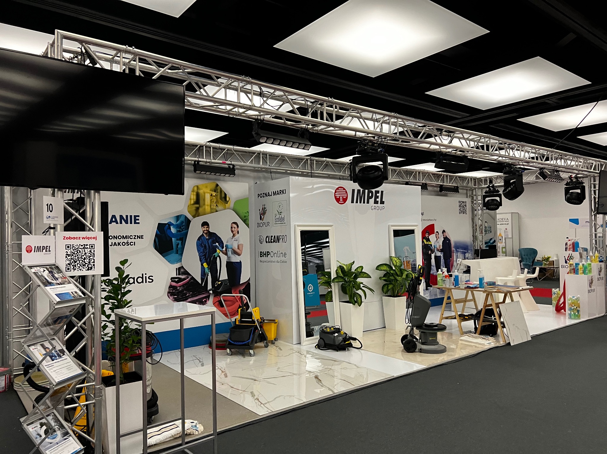 Stoisko targowe firmy oferującej środki czystości i sprzęt do sprzątania, prezentujące maszyny czyszczące, środki chemiczne i materiały reklamowe z logo IMPEL GROUP, BIOPUR, CLEANPRO i BHPOnline.