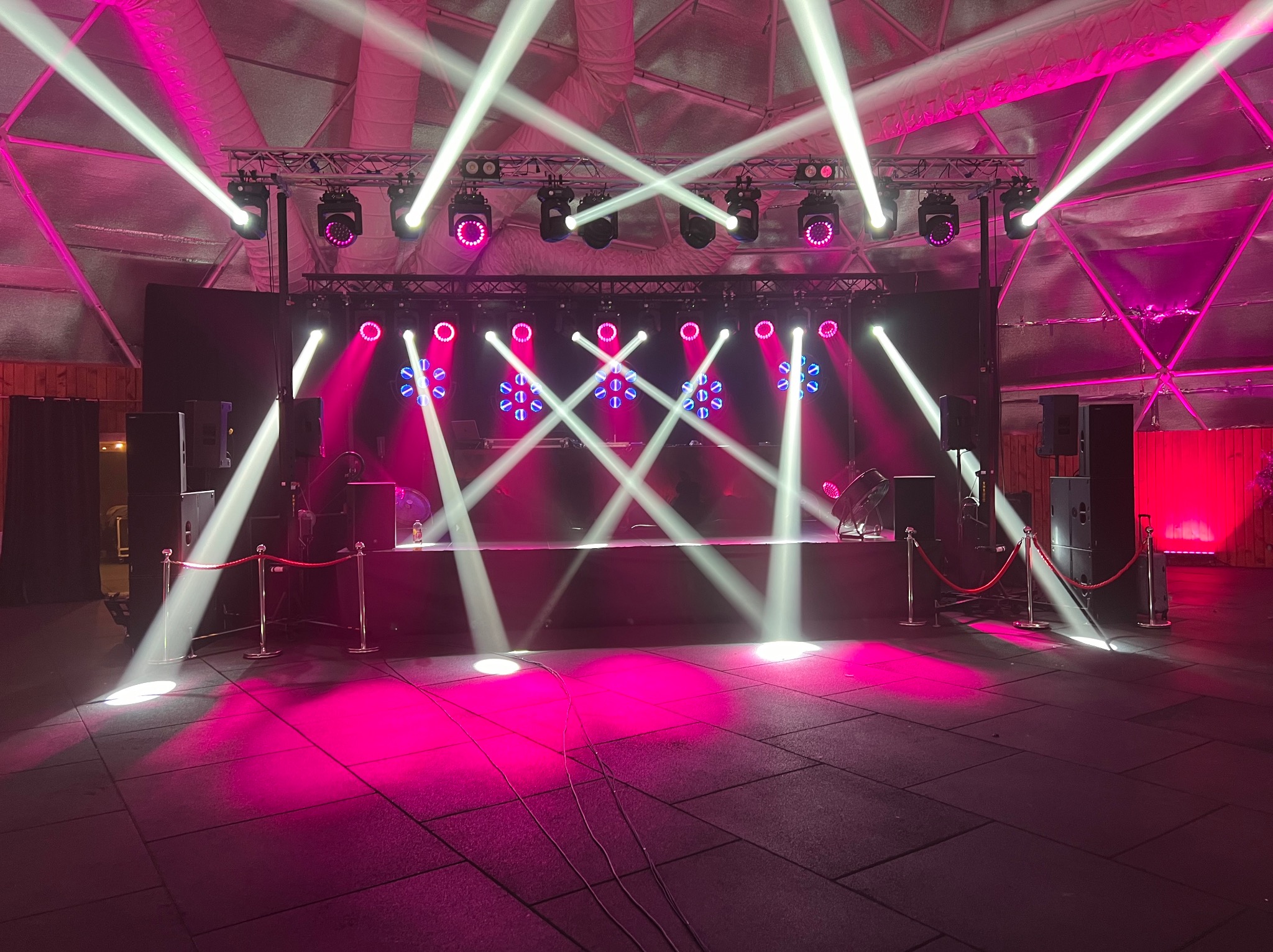 Scena oświetlona białymi i różowymi reflektorami, widoczne kolumny głośnikowe, barierki z czerwonym sznurem, podłoga z szarych płyt, oświetlenie tworzy efektowne krzyżowe wzory.