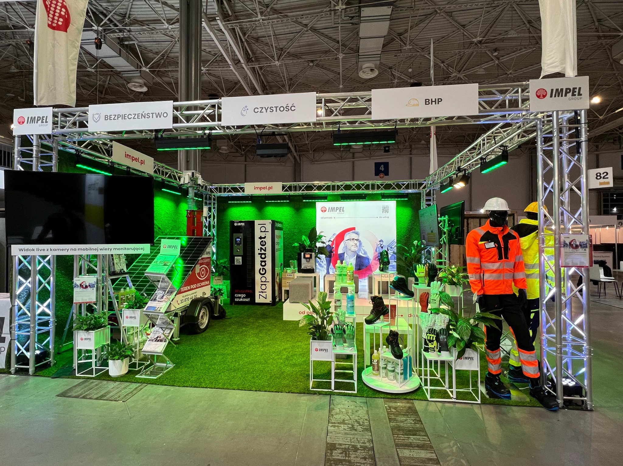 Stoisko firmy Impel na targach, prezentujące usługi z zakresu bezpieczeństwa, czystości i BHP. Ekspozycja zawiera manekiny w odzieży roboczej, system monitoringu wizyjnego na przyczepie, automat...