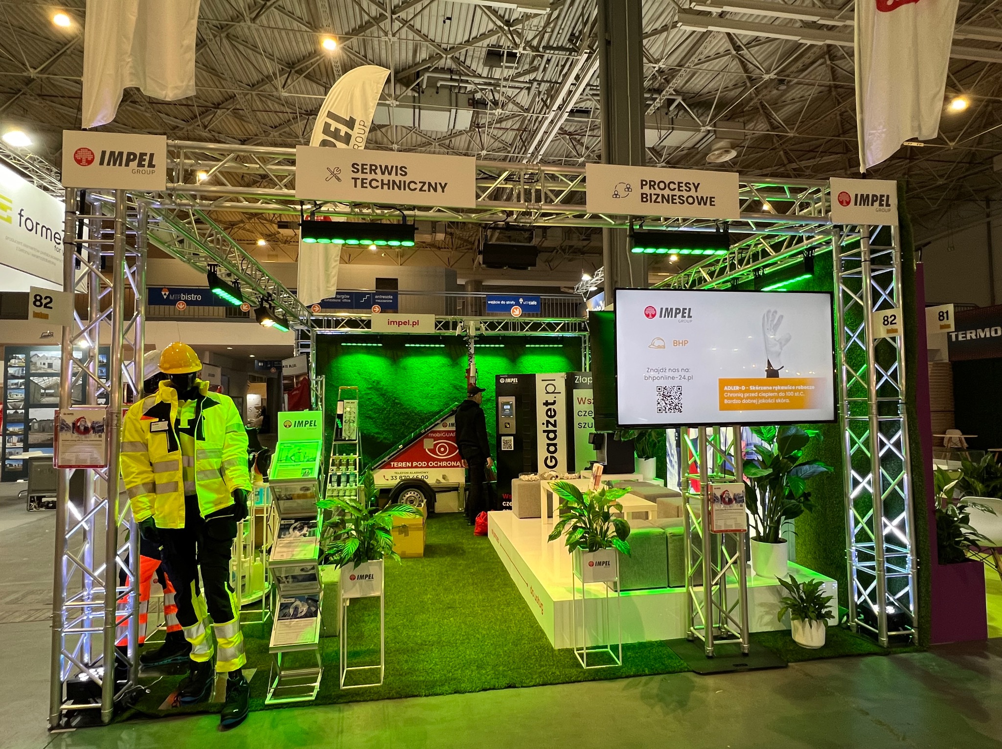 Stoisko firmy Impel Group na targach, z manekinem w odzieży roboczej, ekranem prezentującym ofertę BHP i roślinami doniczkowymi.