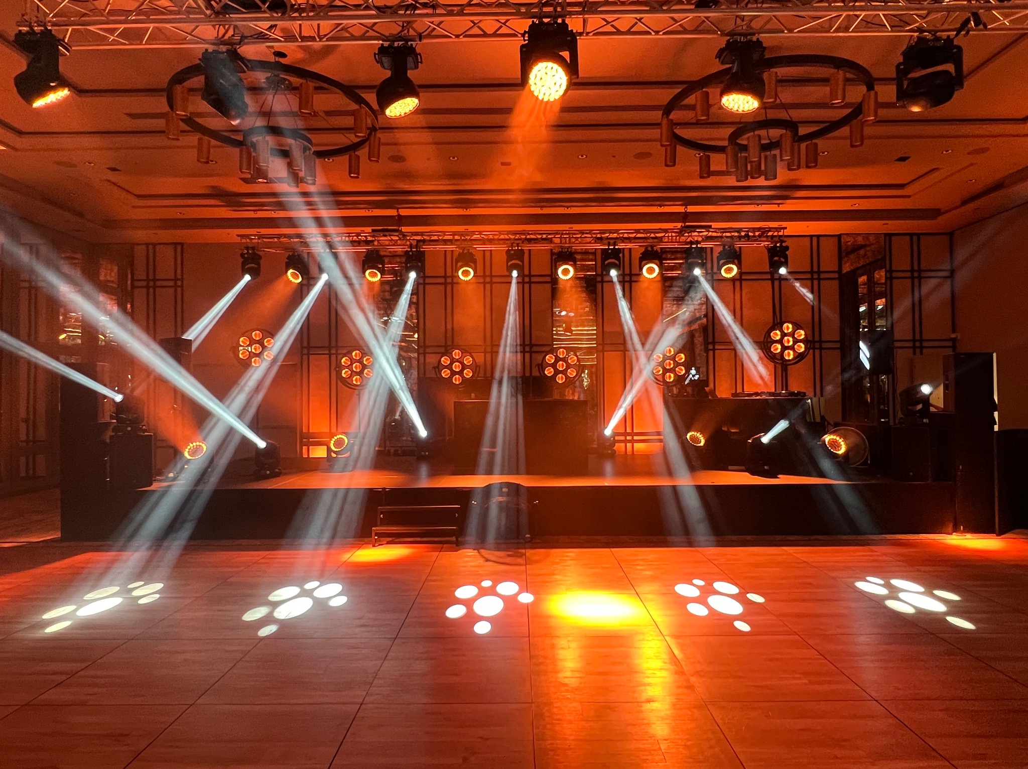 Scena oświetlona reflektorami podczas przygotowań do wydarzenia, widoczne efekty świetlne na parkiecie i sprzęt DJ-ski w tle.