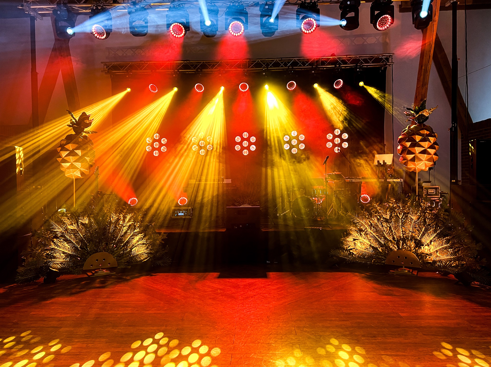 Scena oświetlona reflektorami w kolorach czerwieni i żółci, z dekoracjami w kształcie ananasów po bokach i roślinnością u podstawy, przygotowana na wydarzenie.