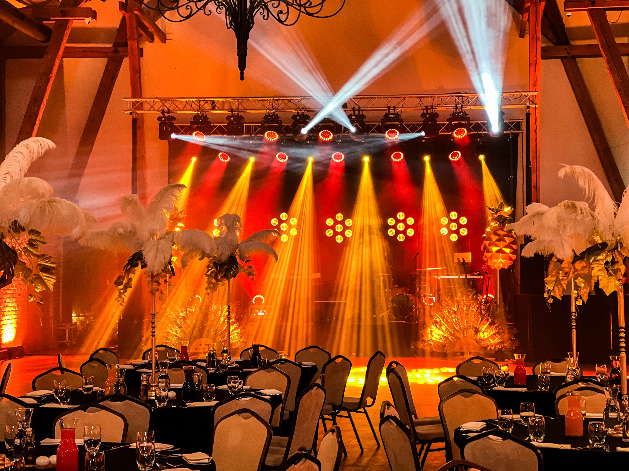 Uroczysta sala balowa z oświetleniem scenicznym, stołami z czarnymi obrusami, białymi piórami w dekoracjach i profesjonalnym nagłośnieniem na scenie