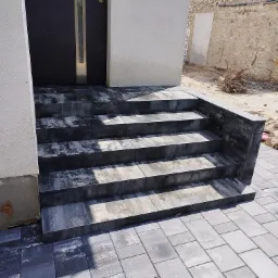 Schody wejściowe z kostki brukowej w odcieniach szarości, prowadzące do ciemnych drzwi z metalową wstawką, z widoczną ścianą w tle w trakcie prac wykończeniowych.