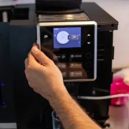 Osoba obsługuje automatyczny ekspres do kawy, wybierając opcję z menu na dotykowym ekranie. Widoczny szklany kubek pod dyszą. W tle wiszą narzędzia.