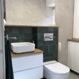 Nowoczesna łazienka z umywalką nablatową na drewnianym blacie, zielonymi płytkami na ścianie i podwieszaną toaletą, uzupełniona o białe szafki i szarą płytkę podłogową.