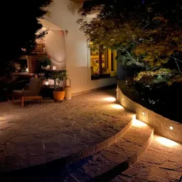 Oświetlony kamienny taras schodkowy nocą, z roślinnością i elementami dekoracyjnymi, w tle budynek z oświetlonym oknem i kominkiem.