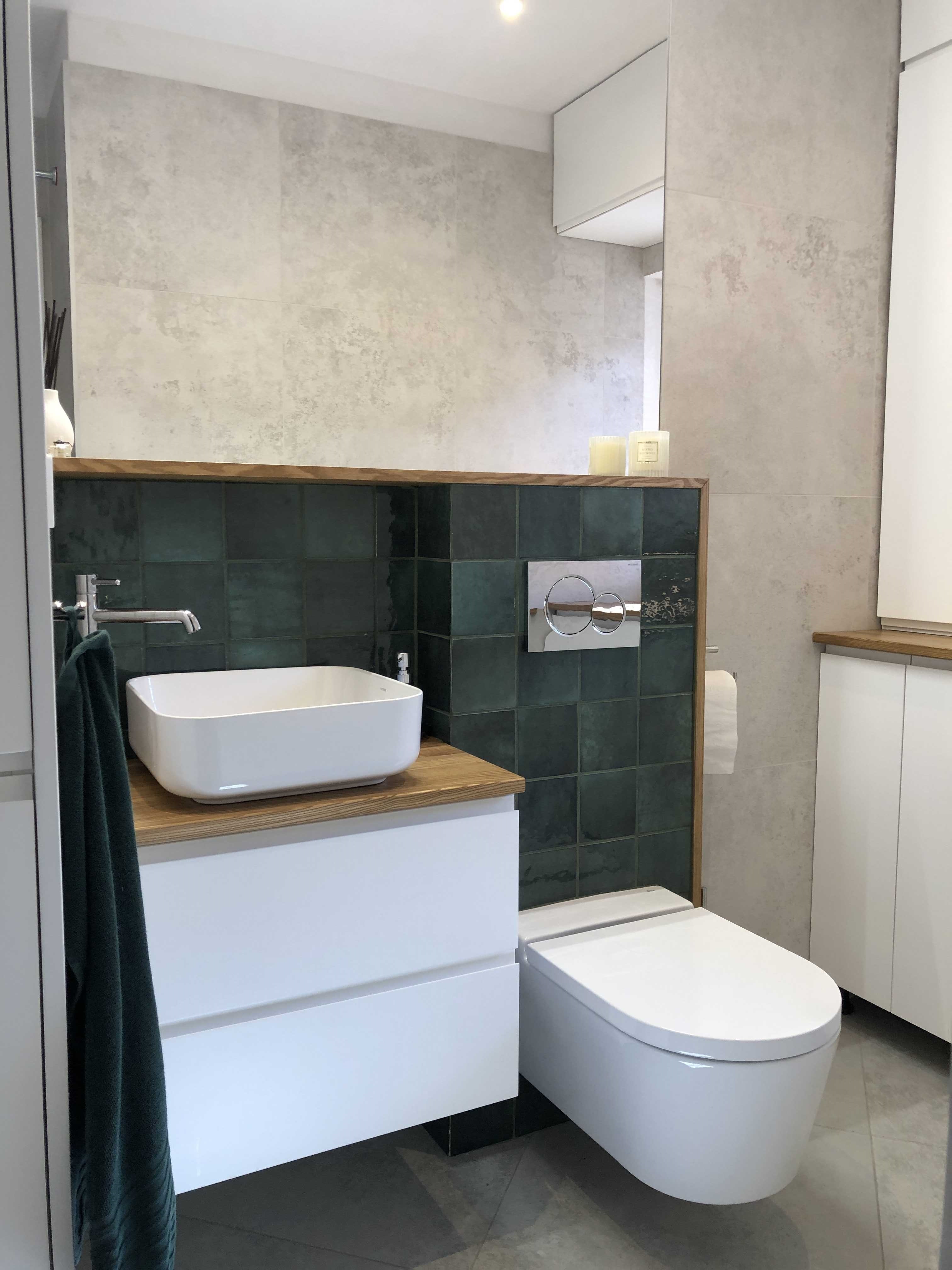 Nowoczesna łazienka z umywalką nablatową na drewnianym blacie, zielonymi płytkami na ścianie i podwieszaną toaletą, uzupełniona o białe szafki i szarą płytkę podłogową.