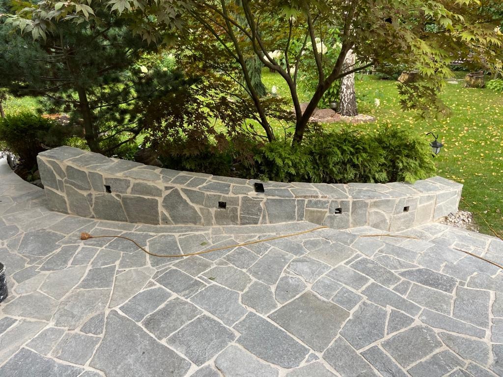 Wykończony taras z nieregularnych płyt kamiennych i niskim murem oporowym, z widocznymi otworami na oświetlenie, otoczony zielenią ogrodu.