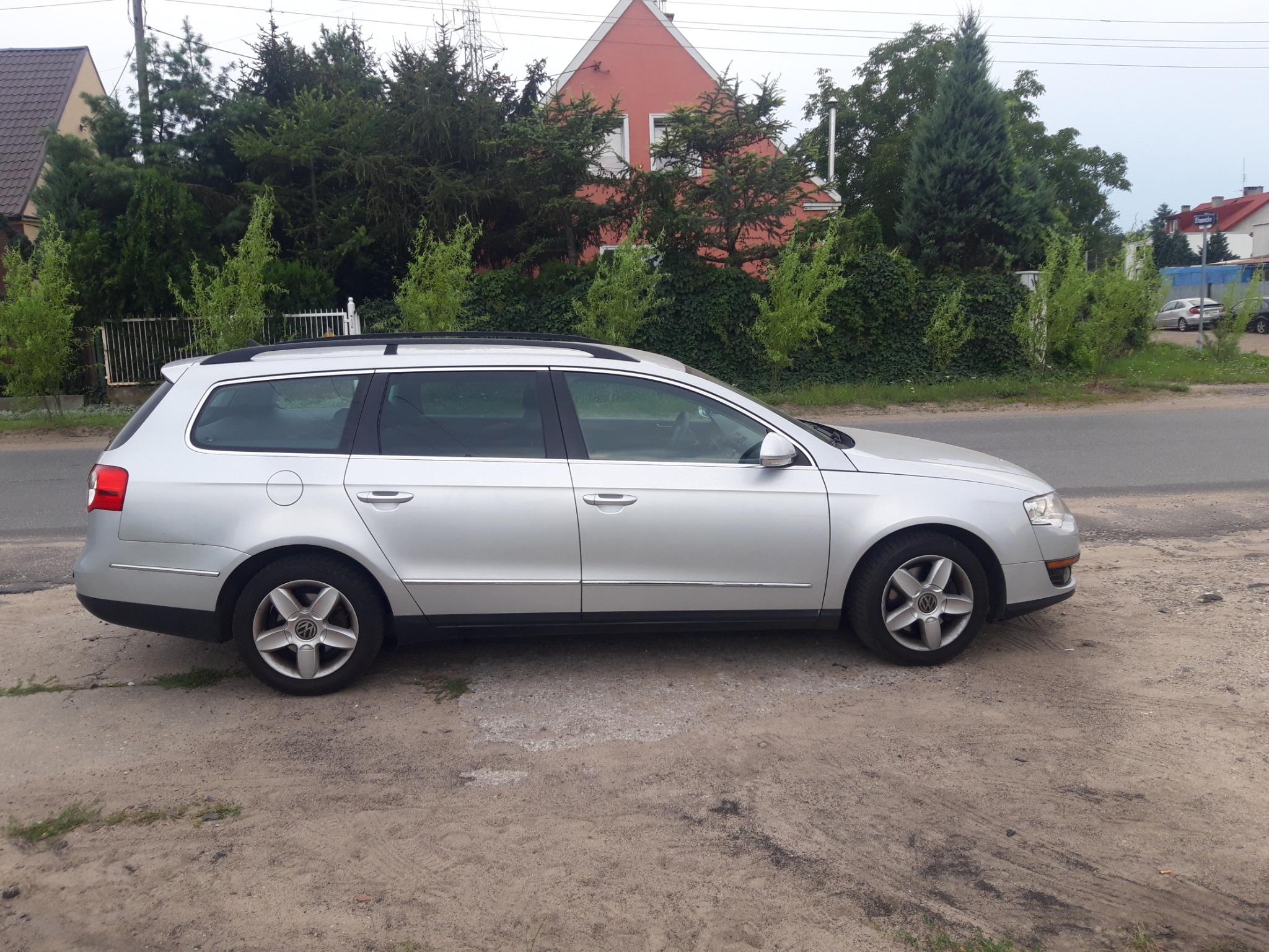 Srebrny samochód kombi zaparkowany przy drodze, z widoczną linią dachu i felgami, na tle zieleni i czerwonego domu.