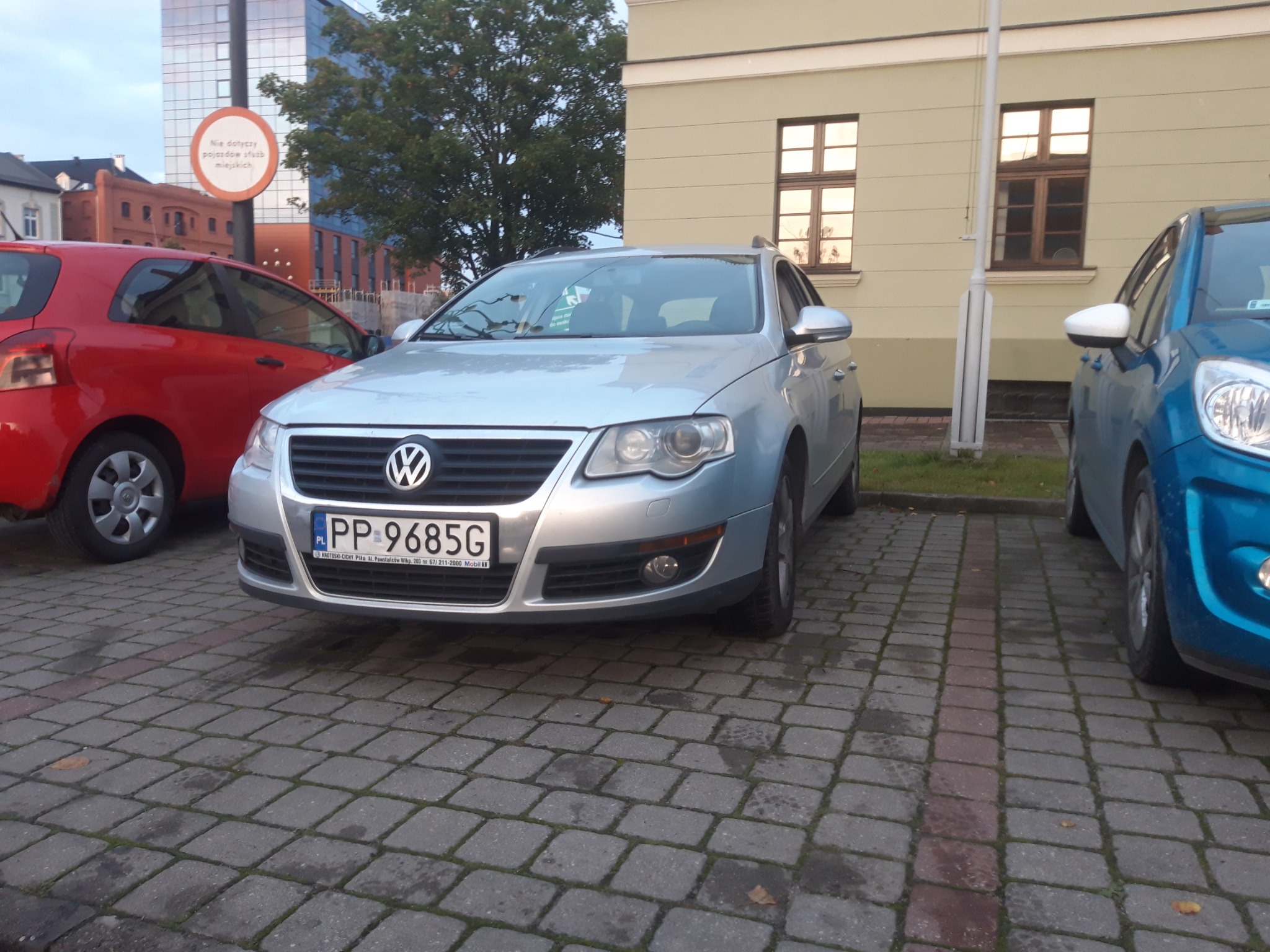 Srebrny Volkswagen Passat zaparkowany na brukowanej ulicy, obok czerwonego i niebieskiego auta, znak drogowy w tle.