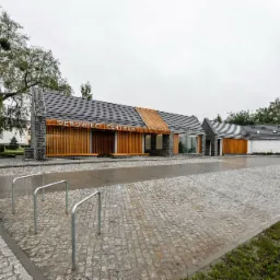 Nowoczesny budynek usługowy Dębowiec Centrum z szarą dachówką i drewnianymi panelami, widok z zewnątrz w pochmurny dzień, na pierwszym planie brukowa nawierzchnia i stojaki na rowery.