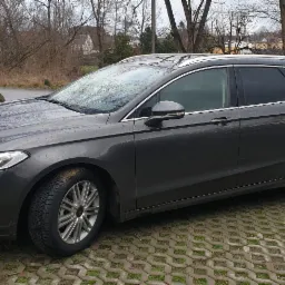 Grafitowy samochód kombi marki Ford, zaparkowany na ażurowym podłożu, z widocznymi kroplami deszczu na karoserii i brudnymi oponami, w tle drzewa bez liści i zabudowania.