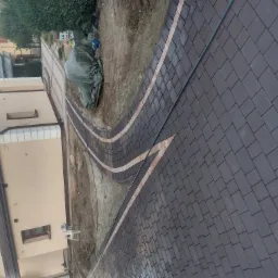 Ukończona ścieżka z ciemnej kostki brukowej z jasnymi obrzeżami, wijąca się obok domu i przygotowanego terenu pod trawnik.