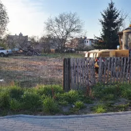 Żółta ładowarka kołowa na placu budowy, widoczna przez drewniany płot, w tle sterta gruzu, samochód typu pick-up i budynki mieszkalne.