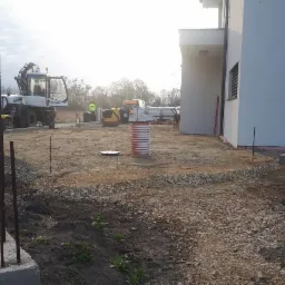 Przygotowanie terenu pod budowę: widoczny fundament z wystającymi prętami, wyrównany teren z gruzem, koparka Mecalac w tle, oraz pracownik w odblaskowej kamizelce przy żółtej mini-koparce.