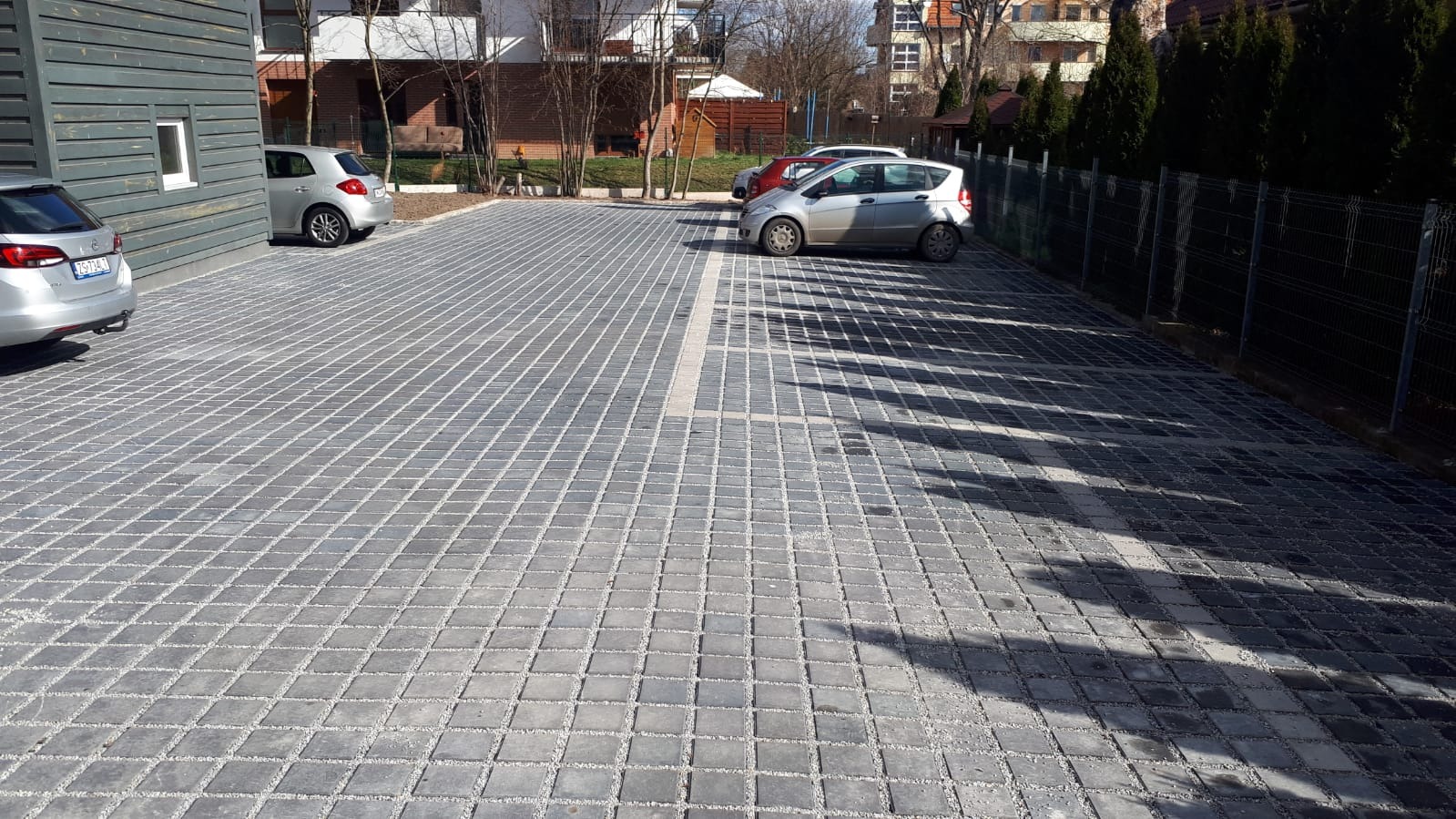 Parking wyłożony szarą kostką brukową z białymi liniami, zaparkowane samochody i cień drzewa.