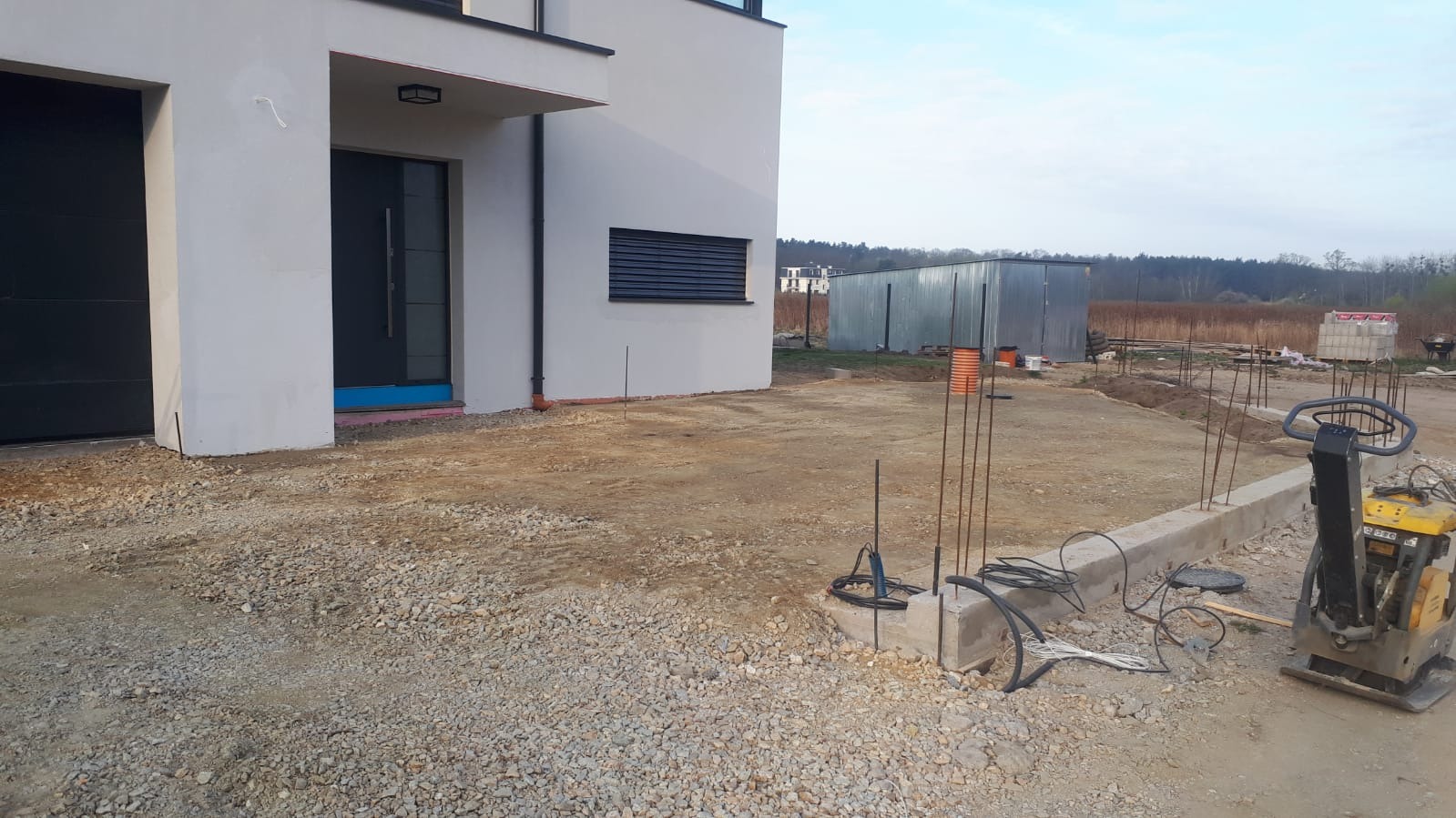 Teren budowy przed nowoczesnym domem z widoczną wibroprasa, przygotowany grunt pod kostkę brukową i zbrojenie fundamentów.
