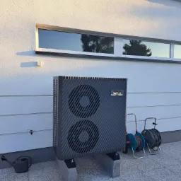 Zewnętrzna jednostka grzewcza Cooper&Hunter na betonowych podstawach, obok szarej konewki i dwóch zwiniętych węży ogrodowych, na tle białej ściany domu z poziomym oknem.