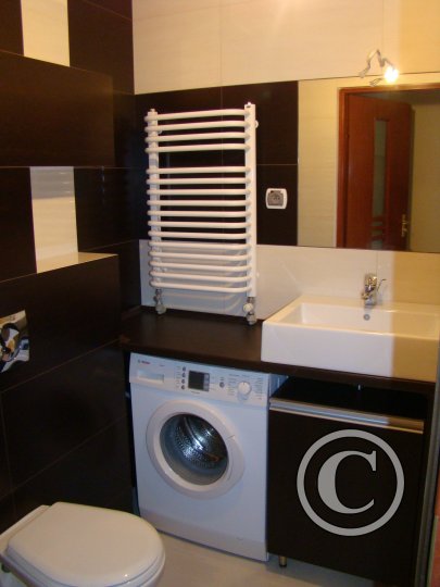 Funkcjonalna łazienka z pralką w zabudowie pod blatem, biała umywalka, grzejnik drabinkowy i płytki w kolorach brązu i bieli.