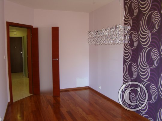 Pomalowane na liliowo wnętrze z drewnianą podłogą, otwartymi drzwiami i ozdobną tapetą w fioletowe wzory na jednej ścianie, oświetlone kryształowym żyrandolem.