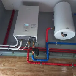 Montaż wiszącego kotła grzewczego Panasonic Aquarea i zasobnika Galmet z izolowanymi rurami w kolorze czerwonym i niebieskim na tle płytek imitujących drewno.