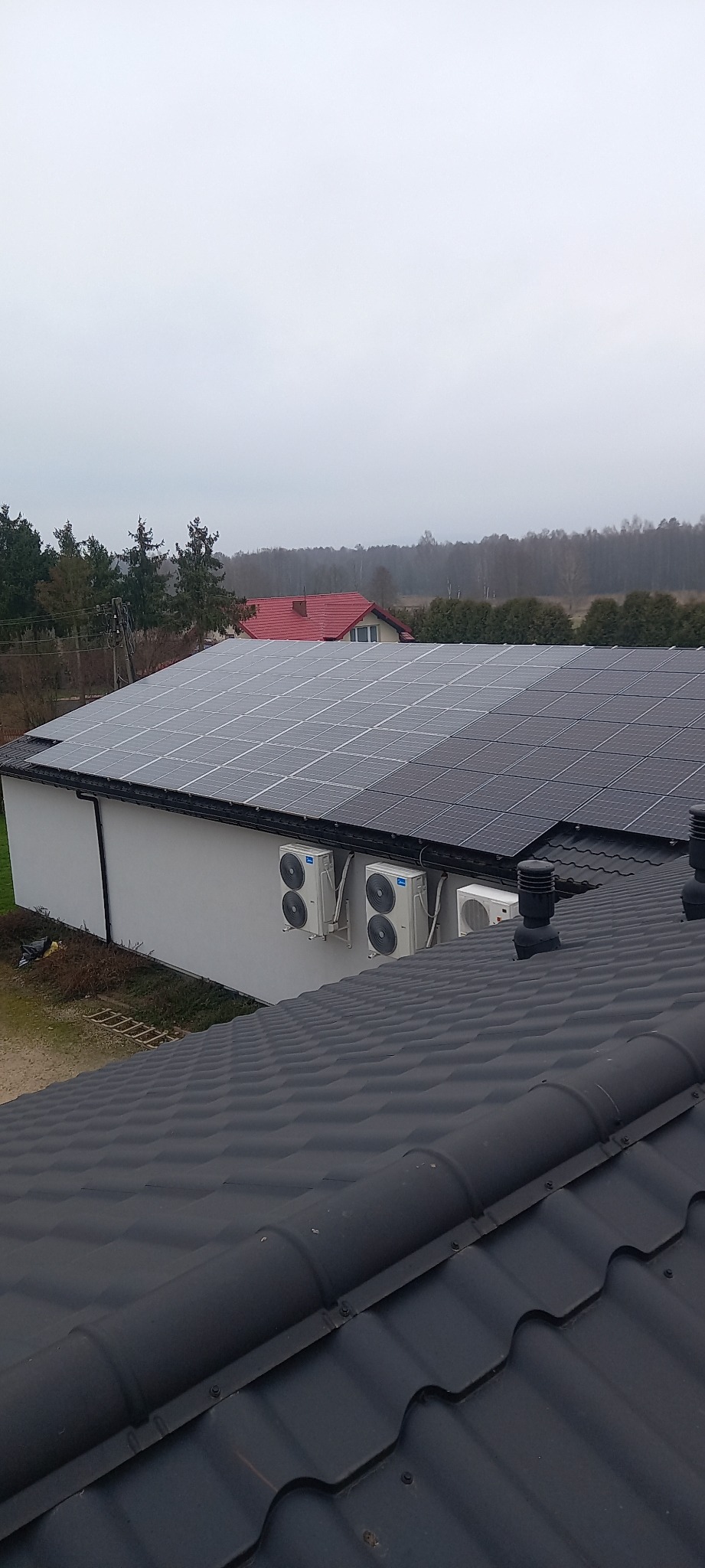 Instalacja fotowoltaiczna na dachu budynku gospodarczego, widoczne panele słoneczne i jednostki klimatyzacyjne na ścianie, w tle las i dom z czerwonym dachem, pochmurne niebo.