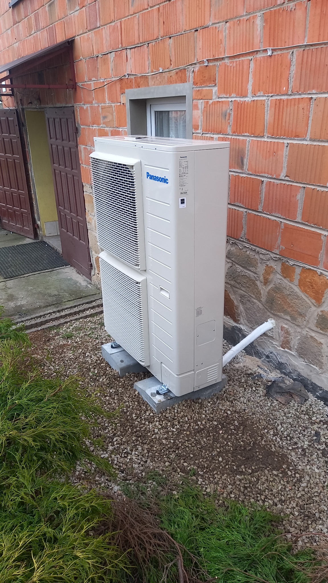 Zewnętrzna jednostka pompy ciepła Panasonic, zamontowana na betonowych podstawach przy ceglanym budynku z widoczną izolacją rury.