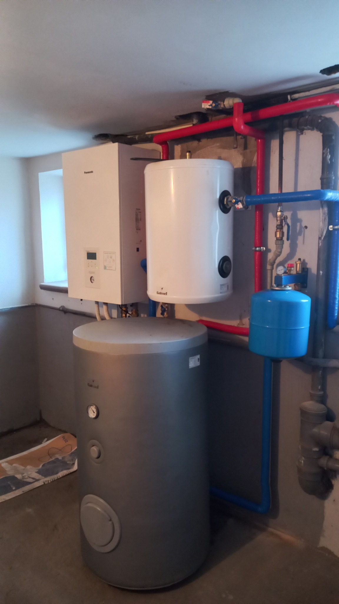 Zestaw urządzeń grzewczych w kotłowni: wiszący biały piec Panasonic, biały zasobnik Galmet i szary, stojący zbiornik buforowy, połączone rurami w kolorach czerwonym i niebieskim.