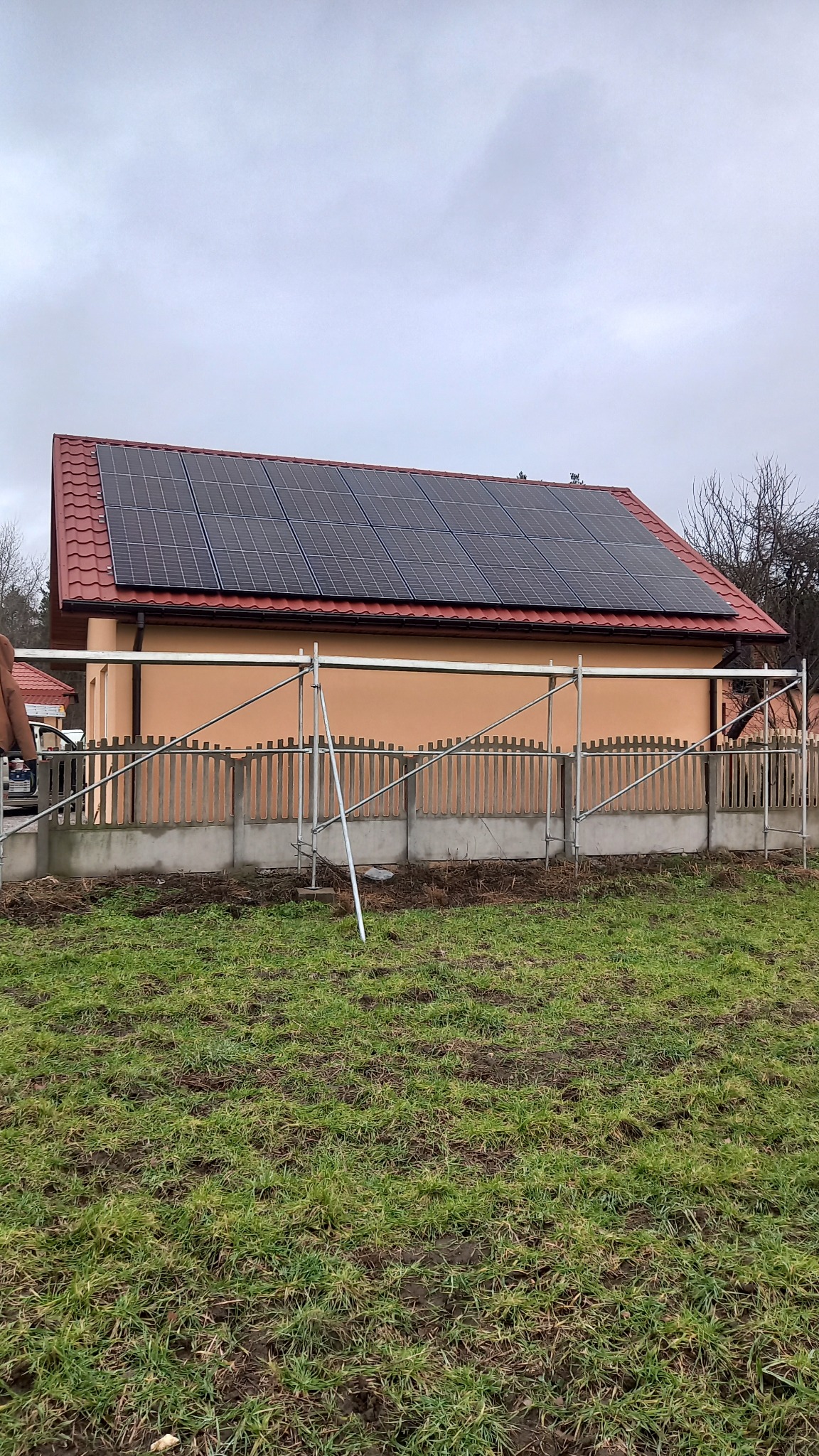 Dom z zainstalowanymi panelami słonecznymi na czerwonym dachu, widoczne rusztowanie przed budynkiem i trawnik.