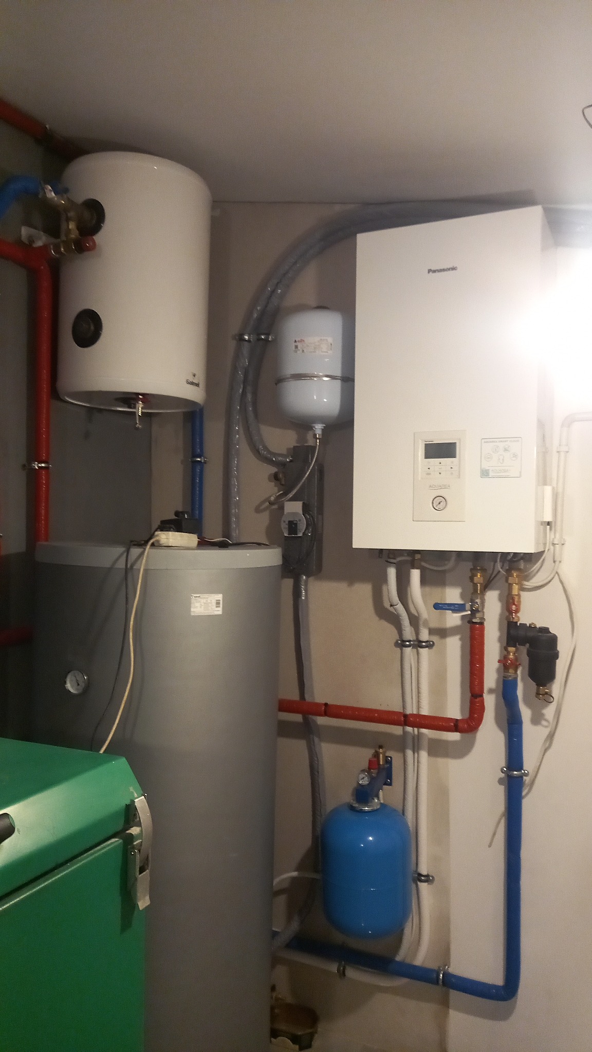 Instalacja pompy ciepła Panasonic Aquarea z zasobnikami CWU, rurami w kolorach czerwonym i niebieskim oraz zielonym zbiornikiem buforowym w tle, w pomieszczeniu technicznym.
