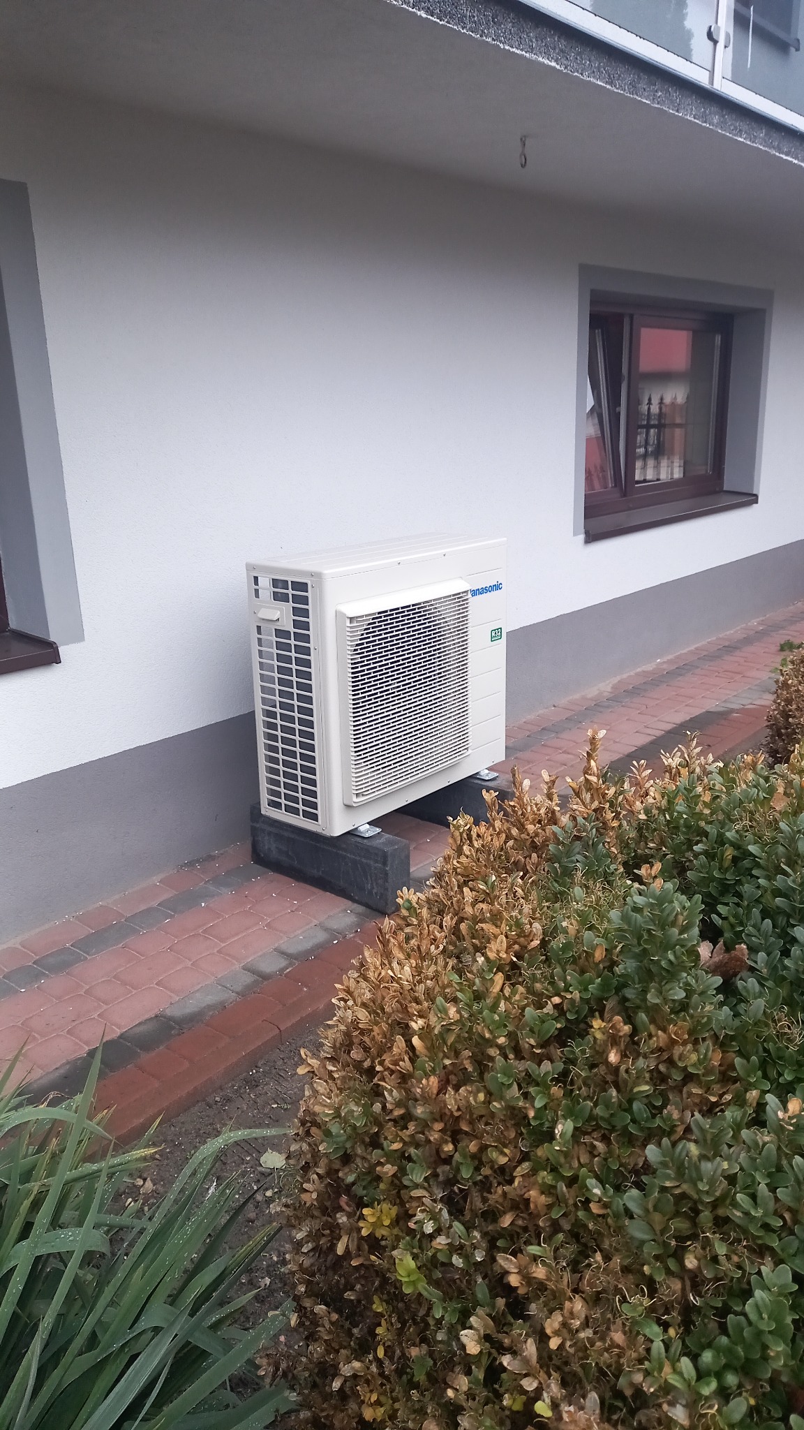 Biała zewnętrzna jednostka pompy ciepła Panasonic zamontowana na betonowych blokach przy ścianie budynku z czerwono-brązową ceglaną ścieżką i częściowo uschniętym żywopłotem.