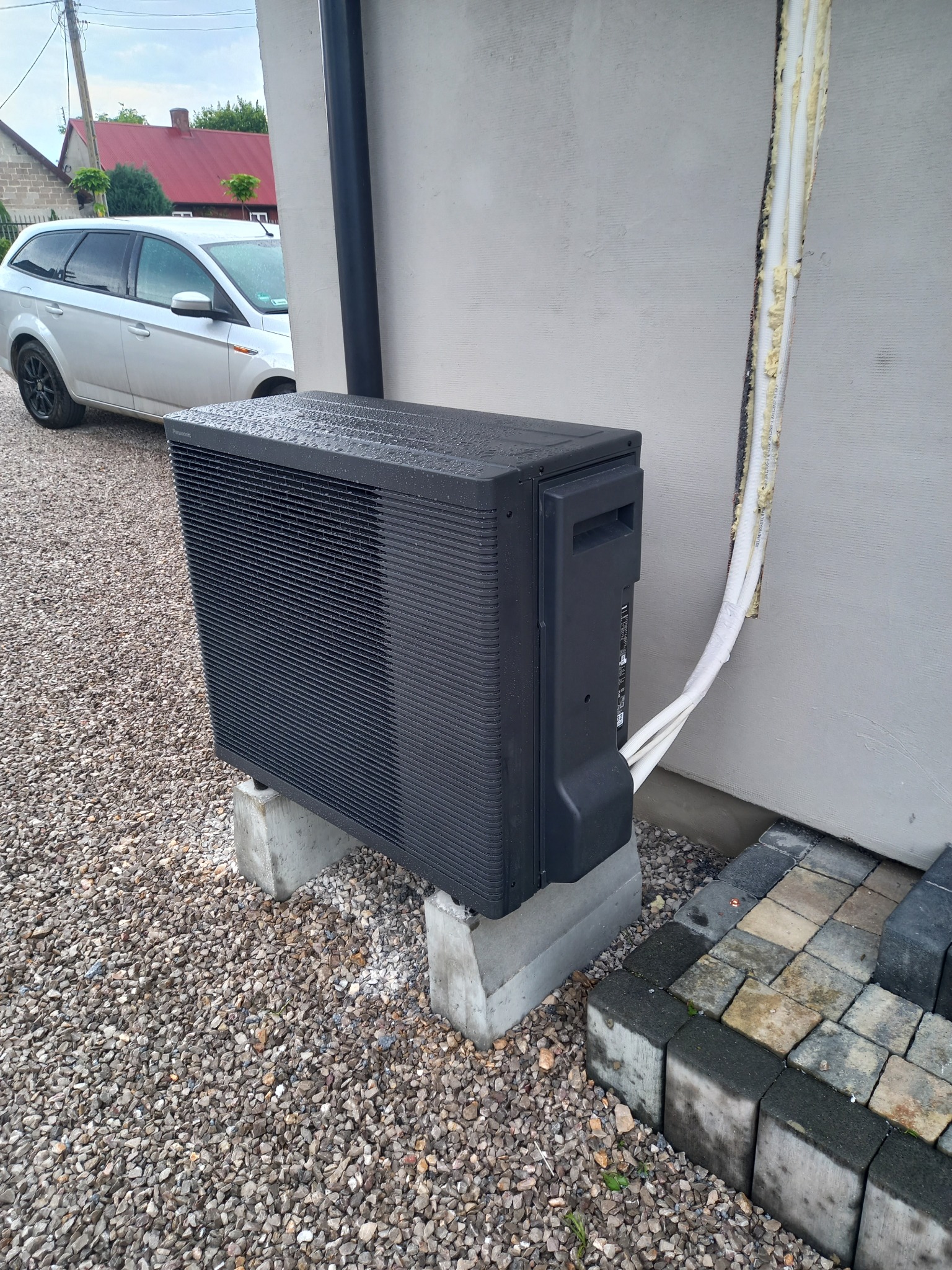 Czarna pompa ciepła, model Panasonic, zamontowana na betonowych podstawach przy jasnoszarym budynku, widoczne rury doprowadzające, na zewnątrz pada deszcz.