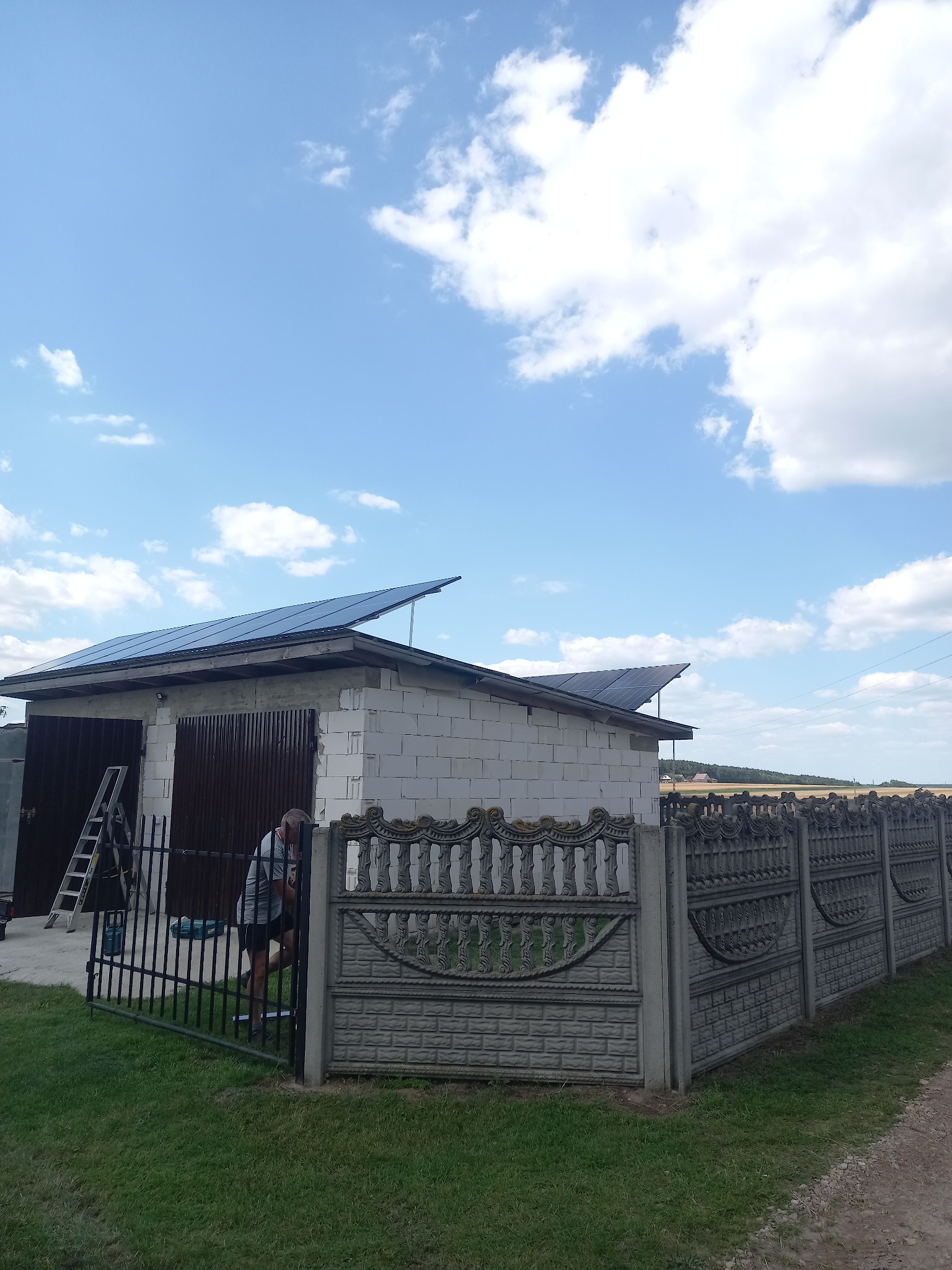 Montaż paneli słonecznych na dachu garażu z białego pustaka, widoczny pracownik przy ogrodzeniu i drabina oparta o ścianę budynku.