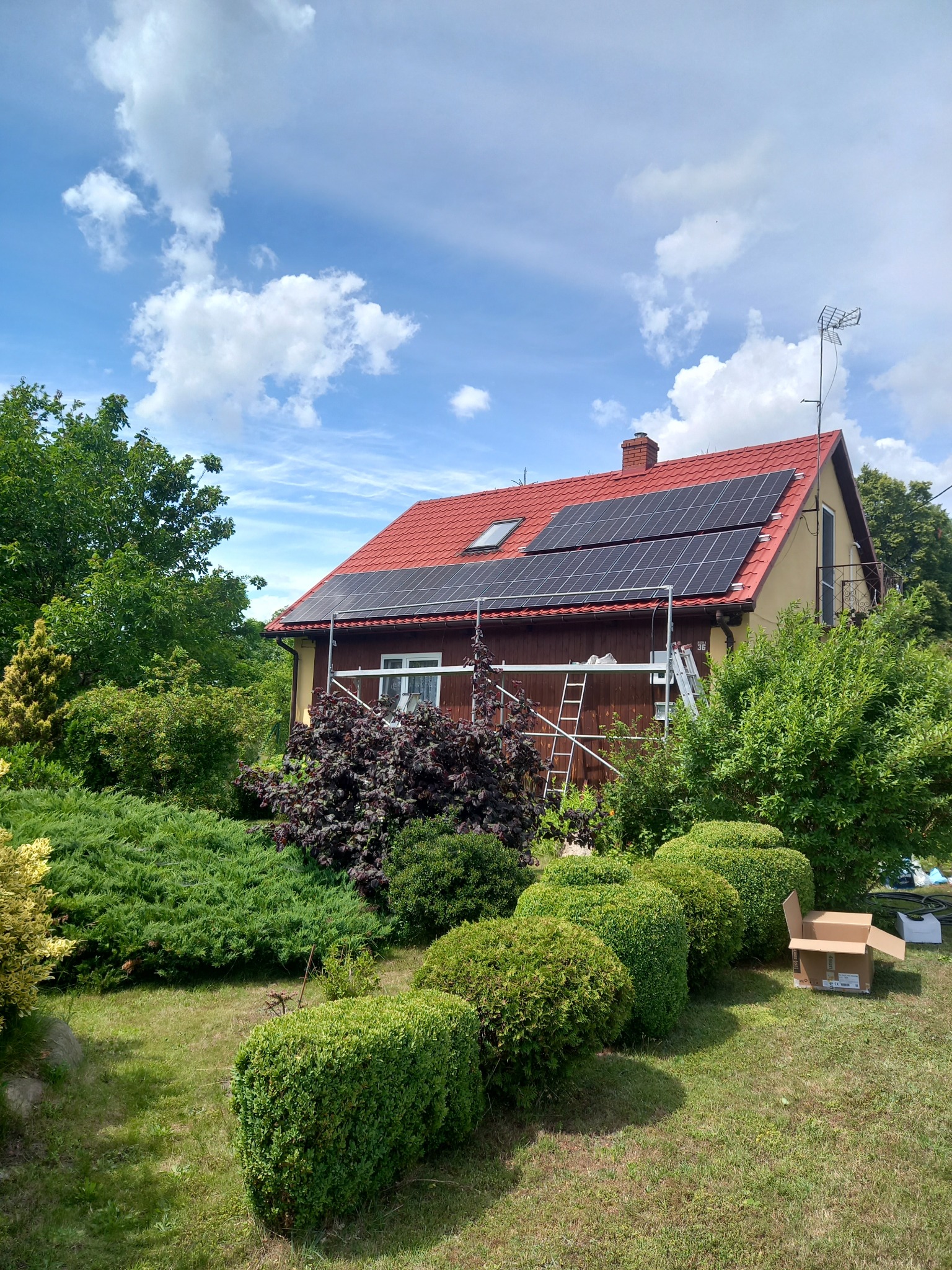 Dom jednorodzinny z czerwoną dachówką i zamontowanymi panelami fotowoltaicznymi, widoczne rusztowanie i drabina, otoczony zielenią w słoneczny dzień.