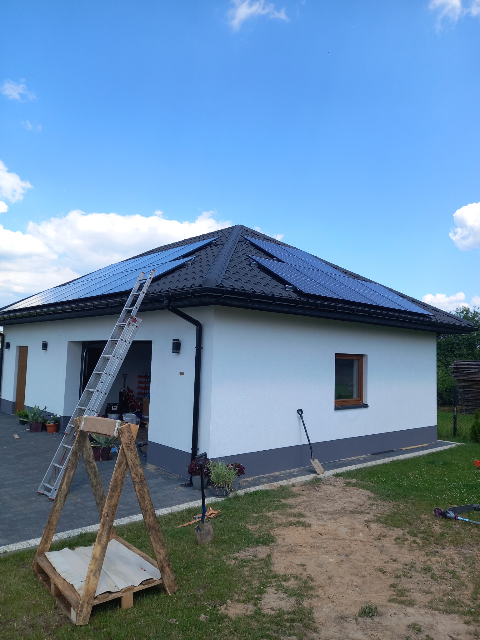 Biały dom jednorodzinny z czarnym dachem pokrytym panelami słonecznymi; aluminiowa drabina oparta o ścianę, drewniany stojak na palecie na trawniku, narzędzia ogrodowe oparte o ścianę.
