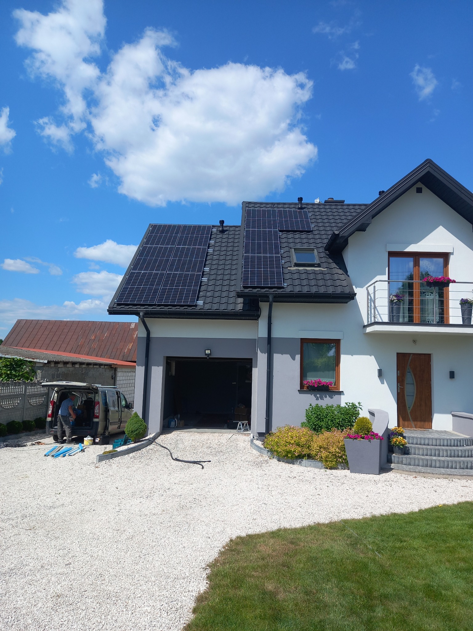 Montaż paneli słonecznych na dachu domu jednorodzinnego; widoczny pracownik przy samochodzie dostawczym z elementami instalacji.