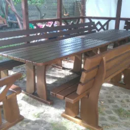 Zestaw mebli ogrodowych z drewna w altanie, składający się z długiego stołu i dopasowanych ławek z oparciami, wykończony w ciemnobrązowym odcieniu.