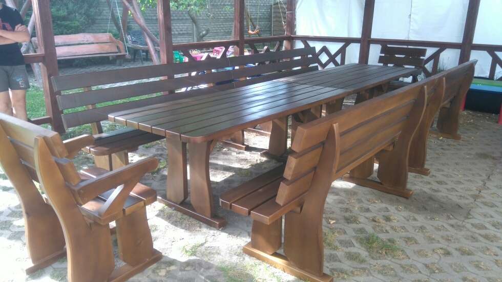 Zestaw mebli ogrodowych z drewna w altanie, składający się z długiego stołu i dopasowanych ławek z oparciami, wykończony w ciemnobrązowym odcieniu.