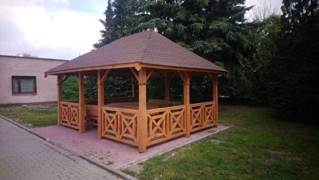 Drewniana altana z brązowym dachem, z dekoracyjnymi panelami z krzyżujących się listew, ustawiona na różowej kostce brukowej, otoczona zielenią i budynkiem w tle.