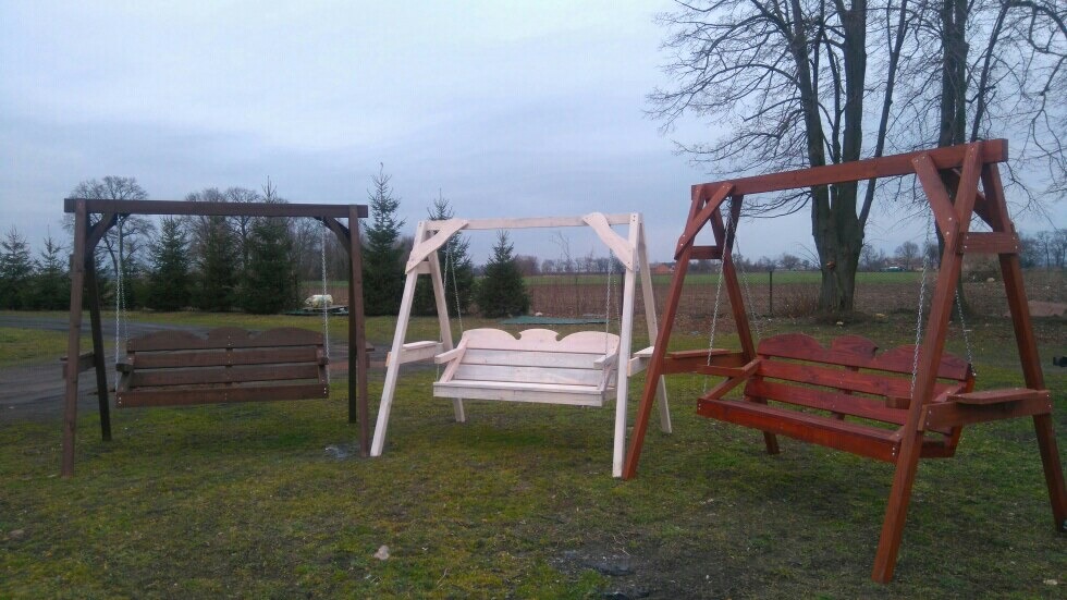 Trzy drewniane huśtawki ogrodowe o różnych odcieniach brązu i naturalnego drewna, ustawione na trawniku, z drzewami w tle.