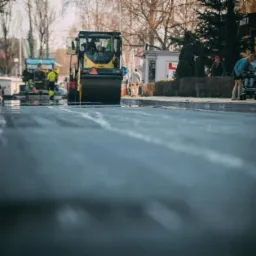 Walec drogowy ubija świeżo wylany asfalt na ulicy w mieście, w tle pracownicy w odblaskowych kamizelkach i przechodnie z wózkiem.